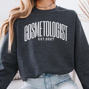 May include: Dark grey heather sweatshirt with the word "COSMETOLOGIST" in white arched lettering, with "EST. 2027" below. The sweatshirt is a crew neck style.