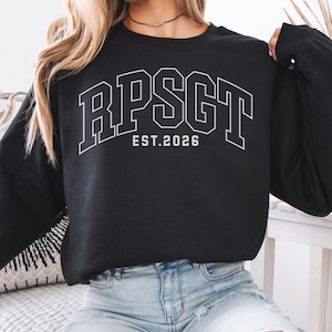 Può includere: Felpa nera girocollo con scritta bianca a blocchi "RPSGT EST. 2026". La felpa è a maniche lunghe e il testo è in un font in stile collegiale. La parola "BLACK" si trova nell'angolo in alto a sinistra.
