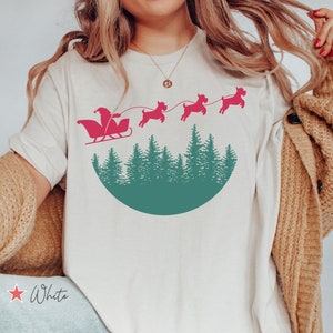May include: White t-shirt featuring a Santa Claus in a sleigh design pulled by dogs, above a green forest silhouette. The text "White" is displayed on the shirt. The Design Boulevard logo is at the bottom.