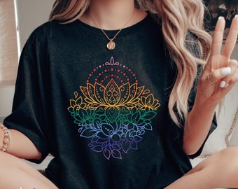 May include: A black t-shirt with a rainbow mandala design. The mandala is a stylized lotus flower with intricate details. The shirt is worn by a person sitting on a bed with a book and a bowl of cereal.