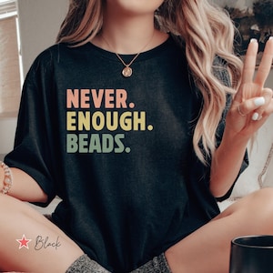 May include: A black t-shirt with the text "NEVER. ENOUGH. BEADS." in rainbow colors. The shirt is worn by a person sitting on a bed with a bowl of cereal and a mug.