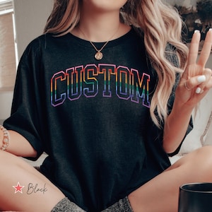 May include: A black t-shirt with the word "CUSTOM" printed in rainbow colors. The shirt is being worn by a person sitting on a bed with a bowl of cereal and a mug.