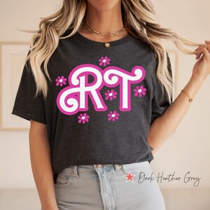 Pu&ograve; includere: Una maglietta grigio scuro melange con un design bianco "RT" circondato da fiori rosa. La maglietta &egrave; indossata da una persona con capelli lunghi biondi e jeans blu chiaro.