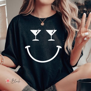 Pode incluir: Uma camiseta preta com um design de rosto sorridente branco. Os olhos do rosto sorridente são copos de martini com azeitonas. O texto "Black" está escrito em vermelho na parte inferior esquerda da imagem.