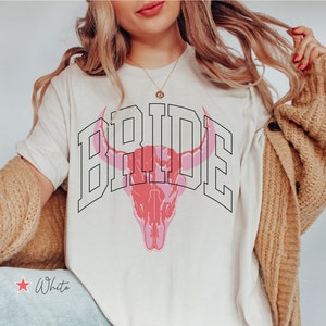 Könnte beinhalten: Weißes T-Shirt mit einem pinkfarbenen Stierkopf-Motiv und dem Wort "BRIDE" in schwarzem Text.