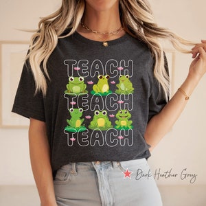 May include: A dark heather grey t-shirt with the word "TEACH" repeated three times in white outlined text. Each word is accompanied by three green frogs sitting on lily pads.