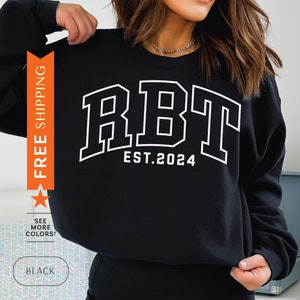 Könnte beinhalten: Schwarzes Sweatshirt mit dem weißen Text "RBT EST. 2024" in einer Schriftart im College-Stil. Das Sweatshirt hat einen Rundhalsausschnitt und lange Ärmel. Das Bild enthält auch die Wörter "SEE MORE COLORS!" und "BLACK".