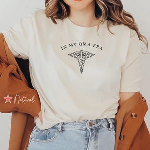 May include: A white t-shirt with the text "IN MY QMA ERA" and a medical caduceus symbol. The shirt is being worn by a person with brown hair and a brown hat.