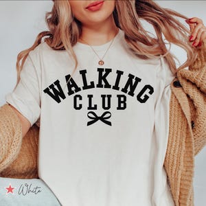 Puede incluir: Camiseta blanca con texto negro que dice "Walking Club" con un lazo negro debajo. La modelo lleva la camiseta con un cárdigan marrón y unos vaqueros azules.