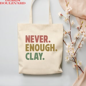 May include: A white canvas tote bag with the text "Never. Enough. Clay." printed in red, yellow, and green. The bag has two handles and is sitting on a light blue surface.