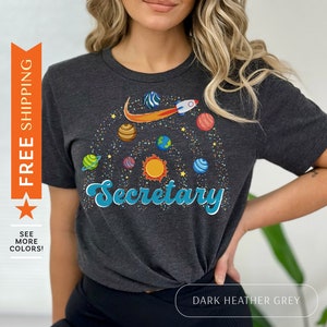 May include: Dark heather grey t-shirt with a graphic of a rocket flying through space with planets and stars. The text "Secretary" is written in a retro font.