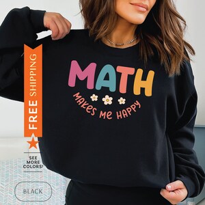 以下が含まれることがあります： カラフルな文字と小さな花のアクセントで「MATH makes me happy」と書かれた黒いスウェットシャツ。