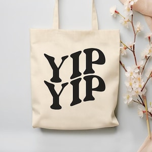 May include: Cream-colored tote bag with the words "YIP YIP" in bold black letters. The bag has two handles and the text "DESIGN BOULEVARD" in red letters. A sprig of white flowers and a peach-colored cloth are in the background.