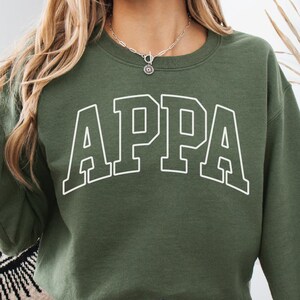 May include: Military green sweatshirt with the word "APPA" in white block letters. The sweatshirt has a crew neck and long sleeves. The text "MILITARY GREEN" is in the upper left corner.