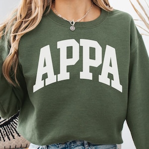 May include: Military green sweatshirt with the word "APPA" in white block letters. The long-sleeved crew neck top is worn with denim shorts. The image also shows a decorative pillow, pumpkins, and a vase.