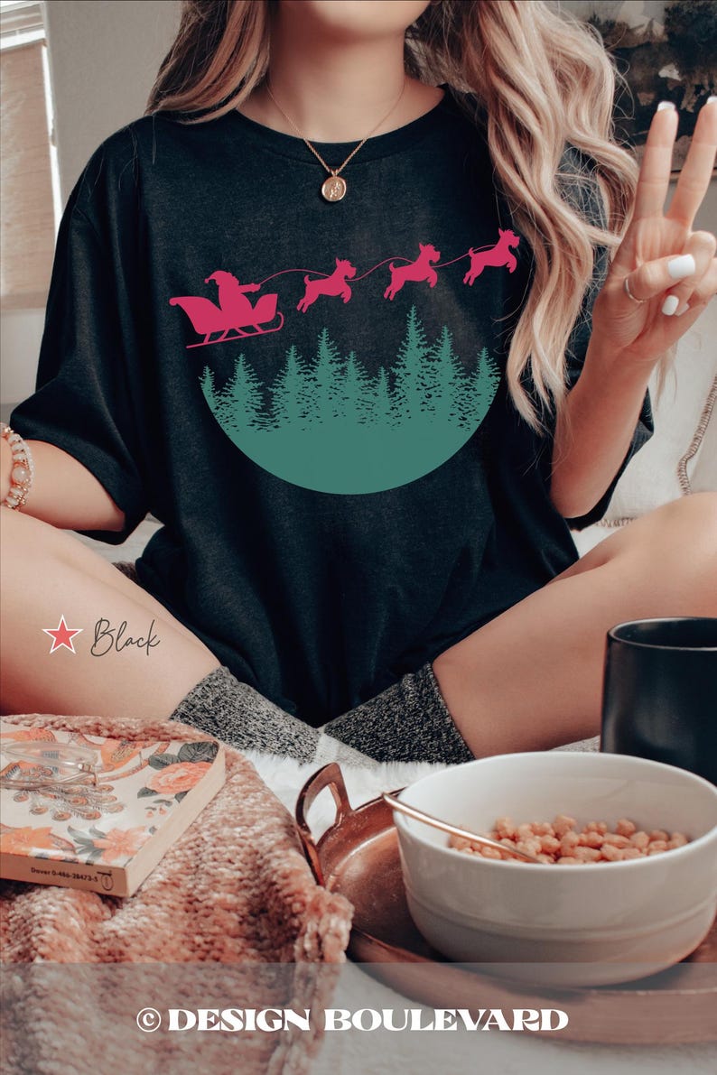 May include: Black t-shirt featuring a pink Santa Claus in a sleigh pulled by pink dogs over a green forest design. The text "Black" is written in a star-shaped outline. The design is from Design Boulevard.