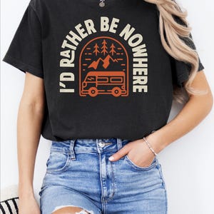May include: Black t-shirt with the text "I'D RATHER BE NOWHERE" in white and orange. The design features a camper van, mountains, and trees. The shirt is worn by a person wearing blue jeans.