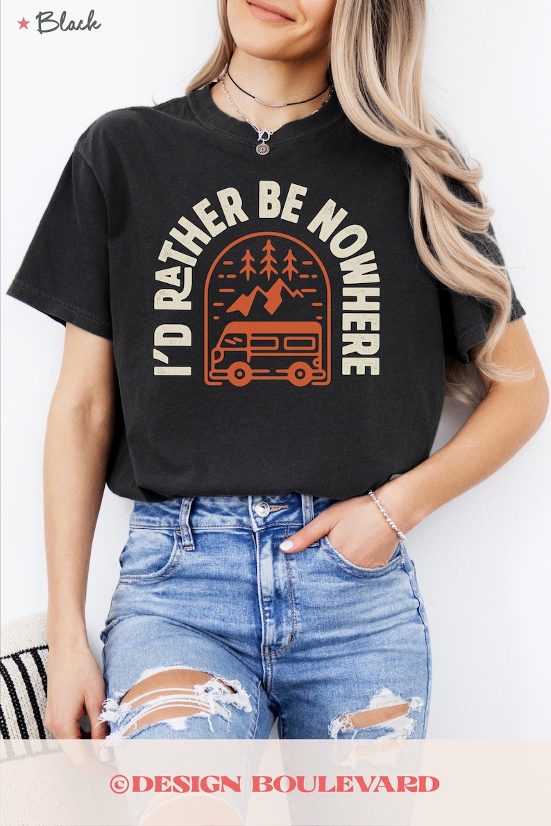May include: Black t-shirt with the text "I'D RATHER BE NOWHERE" in white and orange. The design features a camper van, mountains, and trees. The shirt is worn by a person wearing blue jeans.