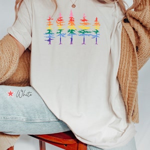 Op de afbeelding: Een cr&egrave;mekleurig T-shirt met korte mouwen en een grafische print van vijf dennenbomen op een rij. Elke boom is gekleurd met horizontale strepen in een regenboogverloop, vari&euml;rend van rood aan de top tot paars aan de onderkant.
