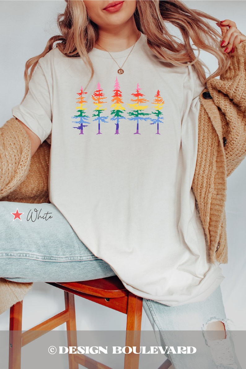 Op de afbeelding: Een cr&egrave;mekleurig T-shirt met korte mouwen en een grafische print van vijf dennenbomen op een rij. Elke boom is gekleurd met horizontale strepen in een regenboogverloop, vari&euml;rend van rood aan de top tot paars aan de onderkant.