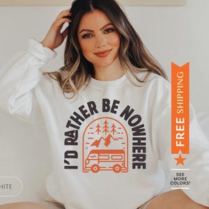 Pode incluir: Camisola branca com um gr&aacute;fico de uma autocaravana em frente a uma cordilheira com &aacute;rvores. O texto "I'd Rather Be Nowhere" est&aacute; em um semic&iacute;rculo em torno da autocaravana.