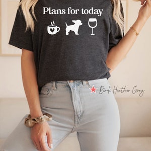 May include: A dark heather grey short-sleeve t-shirt featuring the white text "Plans for today" above three white icons: a coffee mug with a heart, a dog silhouette, and a wine glass. The shirt is paired with light blue distressed denim jeans.