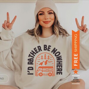 Pode incluir: Uma camisa de moletom bege com o texto "I'd rather be nowhere" em um arco preto com uma van e pinheiros dentro. O texto est&aacute; em laranja e preto.