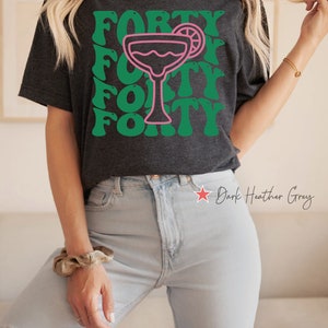 May include: A dark heather grey short-sleeve t-shirt featuring a pink outline of a margarita glass with a lime slice. The word FORTY is printed in a repeating green wavy font pattern behind the glass. The shirt has a crew neckline and short sleeves.
