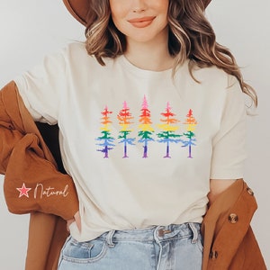 Op de afbeelding: Een cr&egrave;mekleurig T-shirt met korte mouwen en een grafische print van vijf dennenbomen op een rij. Elke boom is gekleurd met horizontale banden in regenboogkleuren, waaronder rood, oranje, geel, groen, blauw en paars. Het T-shirt wordt gedragen met een bruine hoed en een bruine jas.