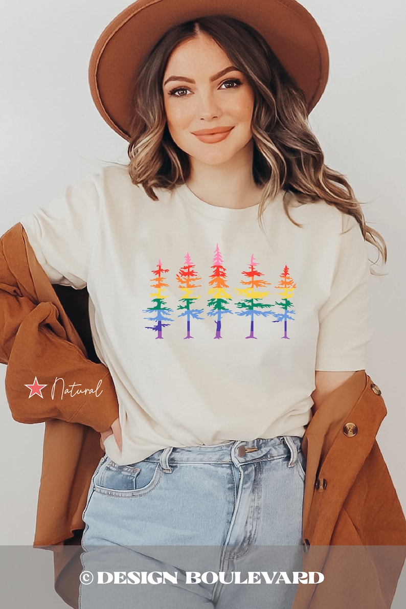 Op de afbeelding: Een cr&egrave;mekleurig T-shirt met korte mouwen en een grafische print van vijf dennenbomen op een rij. Elke boom is gekleurd met horizontale banden in regenboogkleuren, waaronder rood, oranje, geel, groen, blauw en paars. Het T-shirt wordt gedragen met een bruine hoed en een bruine jas.