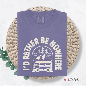 May include: A violet-colored t-shirt with the text "I'D RATHER BE NOWHERE" in a circular design featuring a camper van, mountains, and trees. The shirt is folded on a woven mat. The word "Violet" is at the bottom.