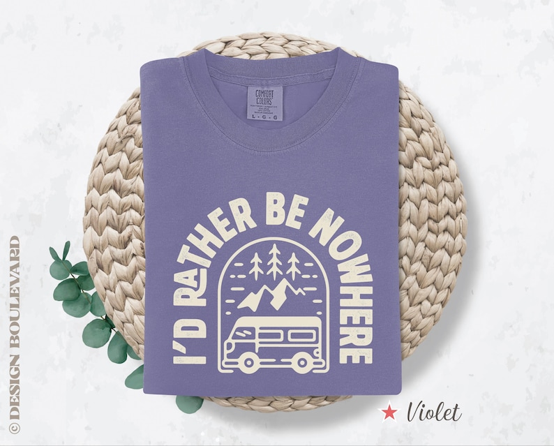 May include: A violet-colored t-shirt with the text "I'D RATHER BE NOWHERE" in a circular design featuring a camper van, mountains, and trees. The shirt is folded on a woven mat. The word "Violet" is at the bottom.