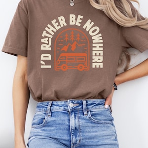 May include: A brown t-shirt with the text "I'D RATHER BE NOWHERE" in cream, featuring an orange camper van graphic. The shirt is worn with blue jeans. The word "Espresso" is in the top left corner.