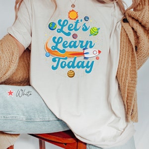 Pode incluir: Camiseta branca com um design gr&aacute;fico de um foguete, planetas e o texto "Let's Learn Today".