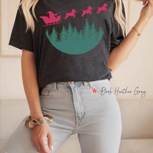 May include: Dark heather grey t-shirt featuring a pink Santa Claus in a sleigh pulled by three pink dogs over a green forest design. The text "Dark Heather Grey" is printed on the shirt. The brand name "DESIGN BOULEVARD" is printed at the bottom.