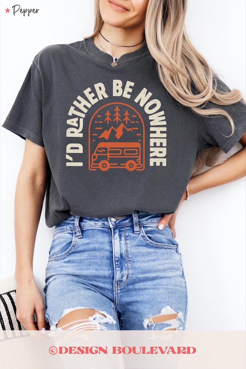 May include: Dark grey t-shirt featuring the text "I'D RATHER BE NOWHERE" in cream, with an orange graphic of a camper van, trees, and mountains. The shirt is a casual style.