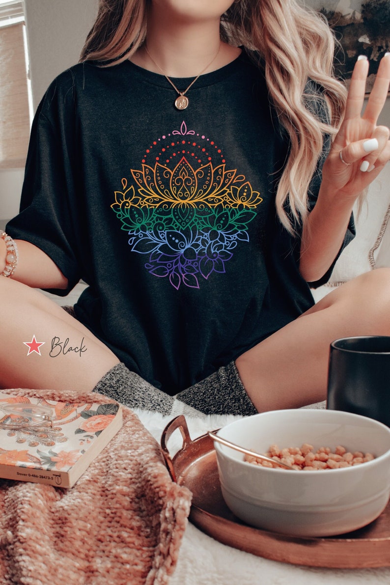 K&ouml;nnte beinhalten: Ein schwarzes T-Shirt mit einem Regenbogen-Mandala-Design. Das Mandala ist eine stilisierte Lotusblume mit filigranen Details. Das Shirt wird von einer Person getragen, die auf einem Bett mit einem Buch und einer Sch&uuml;ssel M&uuml;sli sitzt.