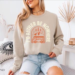 May include: A sand-colored sweatshirt with the text "I'D RATHER BE NOWHERE" in a circular design. The design includes a camper van, trees, and mountains in orange. The sweatshirt is a crew neck style.
