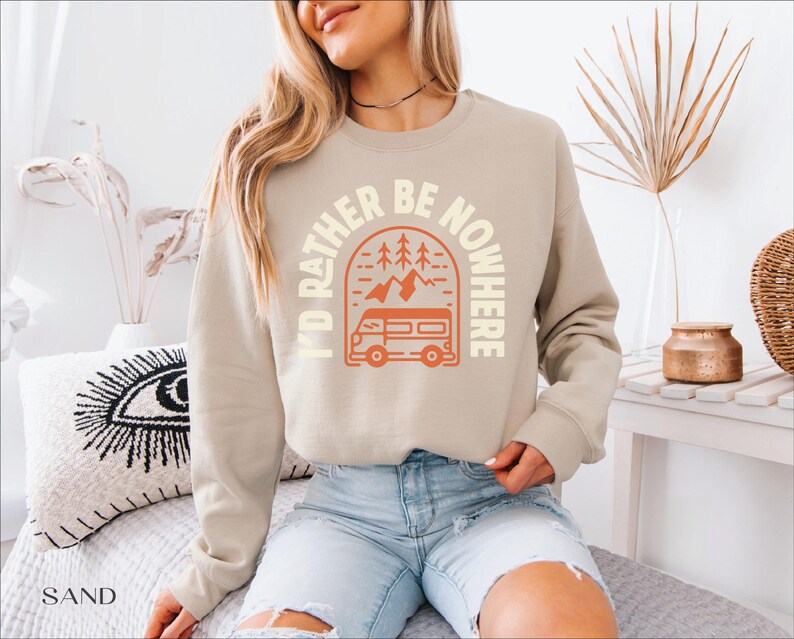 May include: A sand-colored sweatshirt with the text "I'D RATHER BE NOWHERE" in a circular design. The design includes a camper van, trees, and mountains in orange. The sweatshirt is a crew neck style.