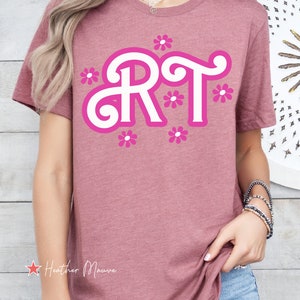 Pu&ograve; includere: Una t-shirt color malva brughiera con una grafica bianca delle lettere "RT" in un font stilizzato con contorni rosa e fiori rosa intorno alle lettere. Il testo "Heather Mauve" &egrave; visibile nell'angolo in basso a sinistra dell'immagine.