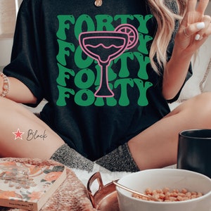 May include: A black t-shirt featuring a graphic print with the word FORTY repeated in a wavy green font. Centered in the design is a pink outline of a margarita glass with a lime wedge garnish. The text is arranged in four rows behind the glass.