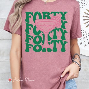 May include: A heather mauve short-sleeve t-shirt featuring a graphic print with the word FORTY repeated in a wavy green font, centered around a pink outline of a margarita glass with a lime slice garnish on the rim of the glass.