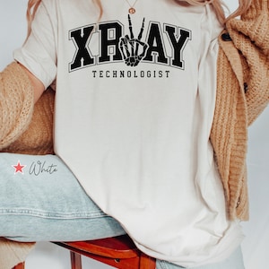 May include: White t-shirt with black text that reads "X-RAY TECHNOLOGIST" and a black skeleton hand making a peace sign.