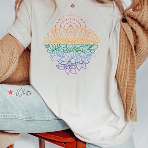 K&ouml;nnte beinhalten: Ein wei&szlig;es T-Shirt mit einem regenbogenfarbenen Lotusbl&uuml;ten-Design. Das Design zeigt ein mandalaartiges Muster mit filigranen Details. Das Shirt wird von einer Person mit langen blonden Haaren getragen.