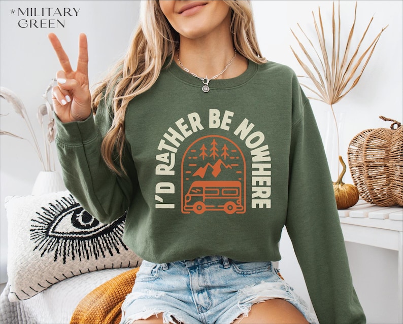 May include: Military green sweatshirt with the text "I'd Rather Be Nowhere" in a white and orange design. The graphic includes a camper van, trees, and mountains. The sweatshirt is paired with blue denim shorts.