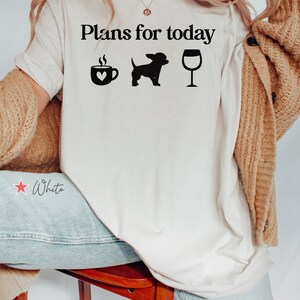 May include: A white t-shirt featuring the black text "Plans for today" above three black icons: a coffee mug with a heart, a small dog silhouette, and a wine glass. The shirt is worn with a tan cardigan and light blue denim jeans.
