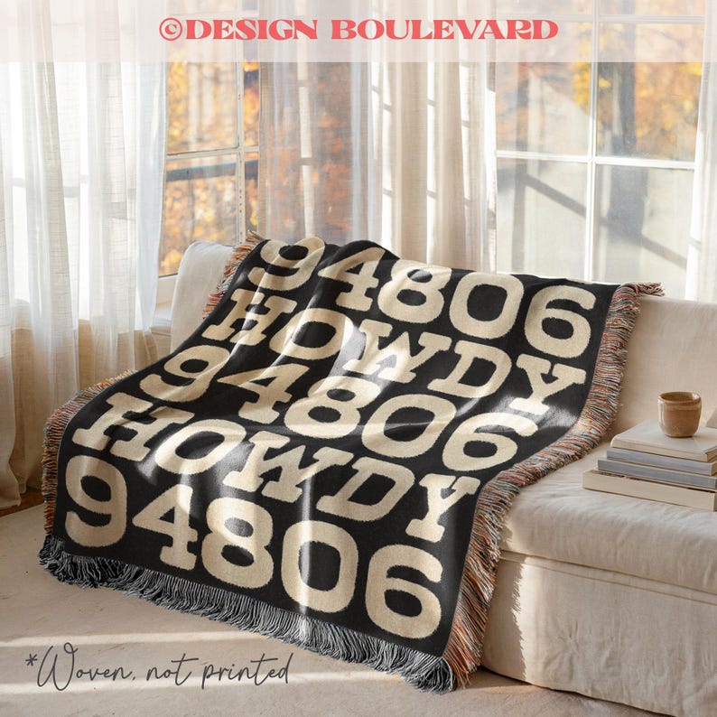 Op de afbeelding: Een zwarte geweven plaid met de herhalende cr&egrave;mekleurige tekst "94806" en "HOWDY" in een vetgedrukt bloklettertype. De deken heeft een rand met franjes en een gestructureerde geweven constructie, met een herhalend patroon over het oppervlak.