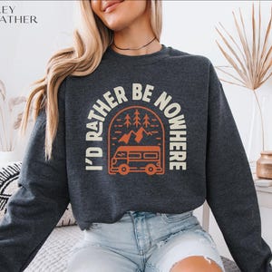 May include: Grey heather sweatshirt with the text "I'd Rather Be Nowhere" in a circular design. The design includes a camper van, trees, and mountains in orange and white. The sweatshirt is a crew neck.