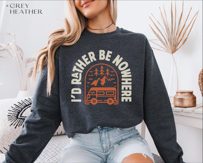May include: Grey heather sweatshirt with the text "I'd Rather Be Nowhere" in a circular design. The design includes a camper van, trees, and mountains in orange and white. The sweatshirt is a crew neck.
