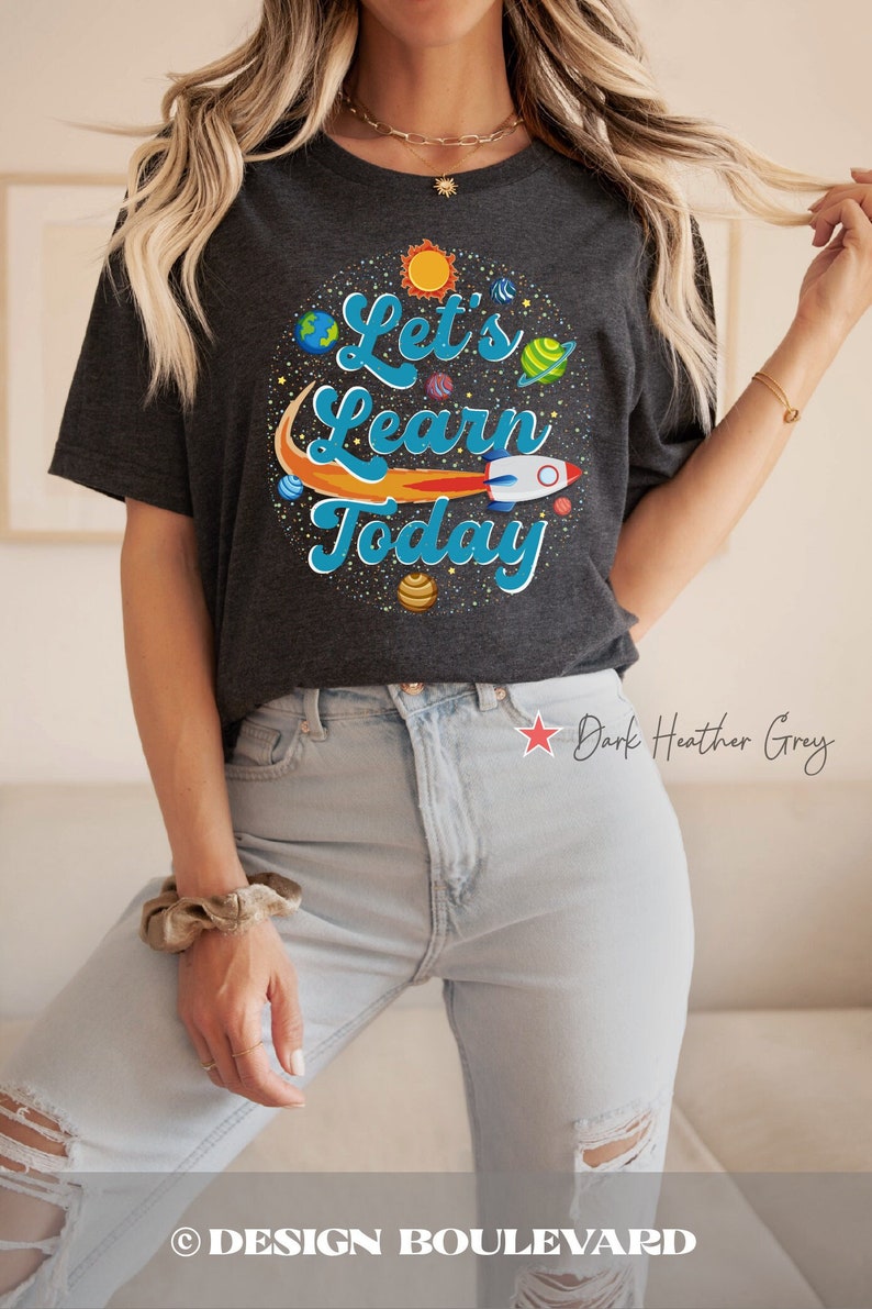 Pode incluir: Uma camiseta cinza escuro mesclada com um gr&aacute;fico de tem&aacute;tica espacial que diz "Let's Learn Today". O gr&aacute;fico inclui um foguete, planetas e estrelas.